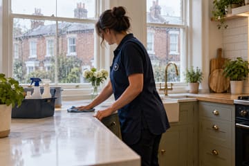 A cleaner wiping down a marble kitchen worktop beside a caddy of cleaning sprays, with rain on the window.