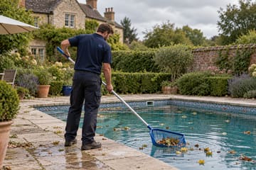 A pool technician skimming leaves from an outdoor pool with a net, in front of a stone house.