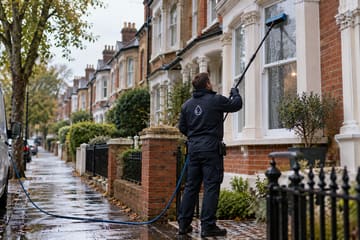 A window cleaner using a water-fed pole on a bay window along a wet terraced street, with a van parked at the kerb.